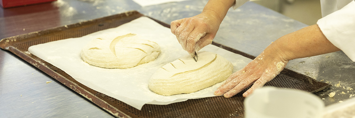 Floured hands use a razor to score curved lines in loaf-shaped dough before baking